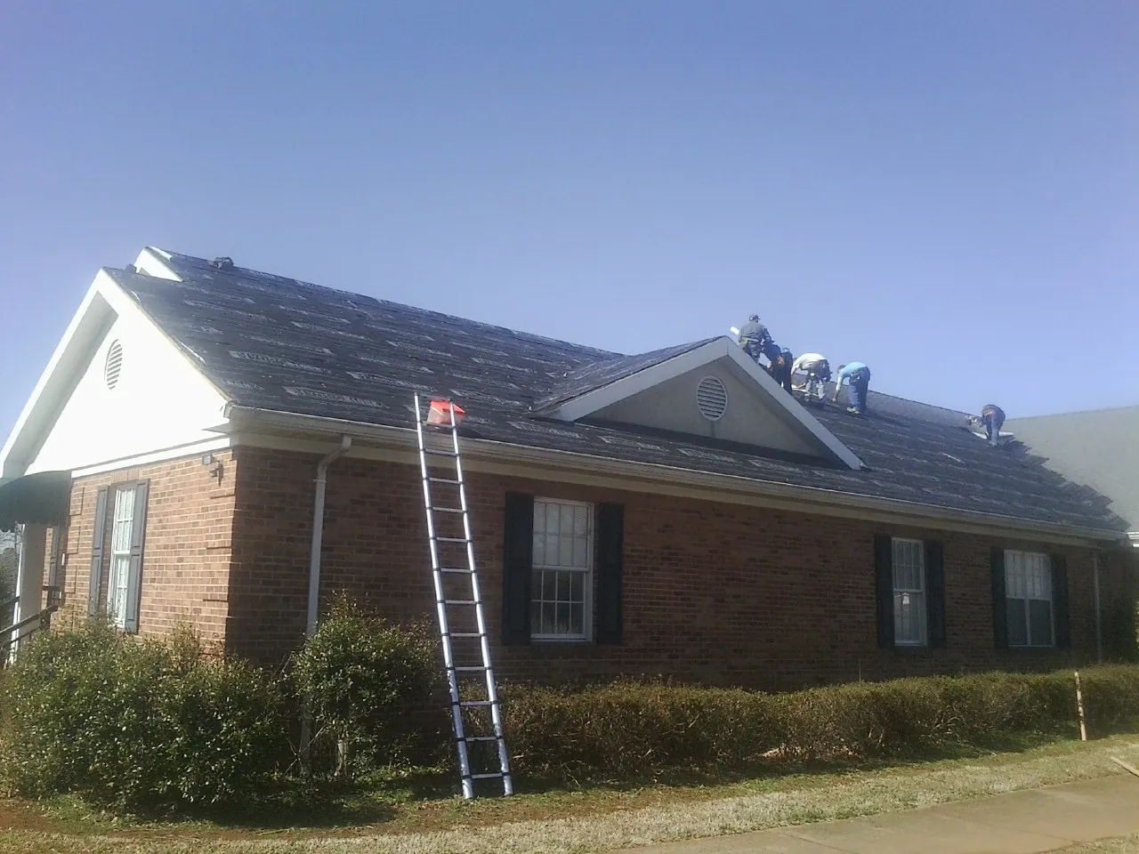 Crew tearing shingles off a brick ranch roof with a ladder set — Metal Roof Repair work by our crew in Collettsville