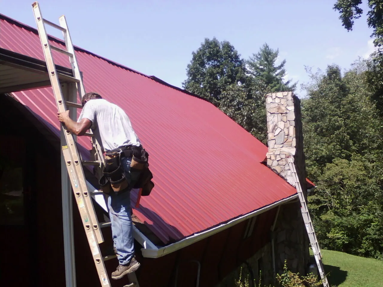 Roofer on a ladder beside a red metal roof and stone chimney — Soffit & Fascia Repair by Roofing Brothers in Collettsville